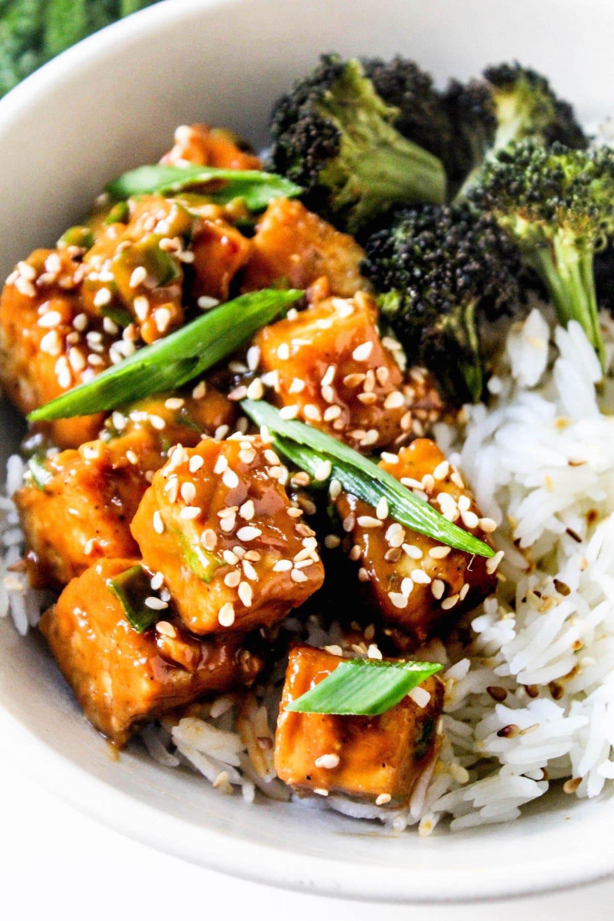 Peanut sauce tofu cubes sit on a bowl of white rice with a side of broccoli inside a white bowl.