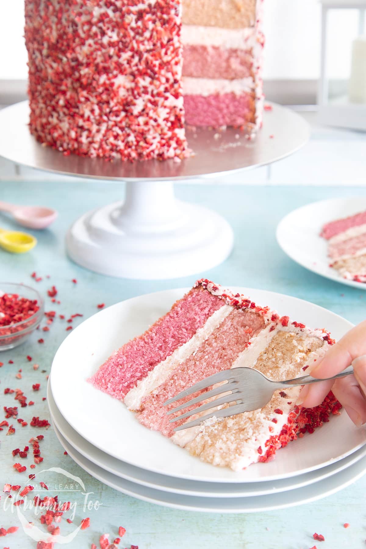 A slice of pink ombre cake on a stack of small white plates. The cake has three layers, each an increasingly intense shade of pink. The sponges are layered with pale pink frosting and the outside is decorated with freeze dried strawberry pieces. A hand holds a fork, delving into the cake.