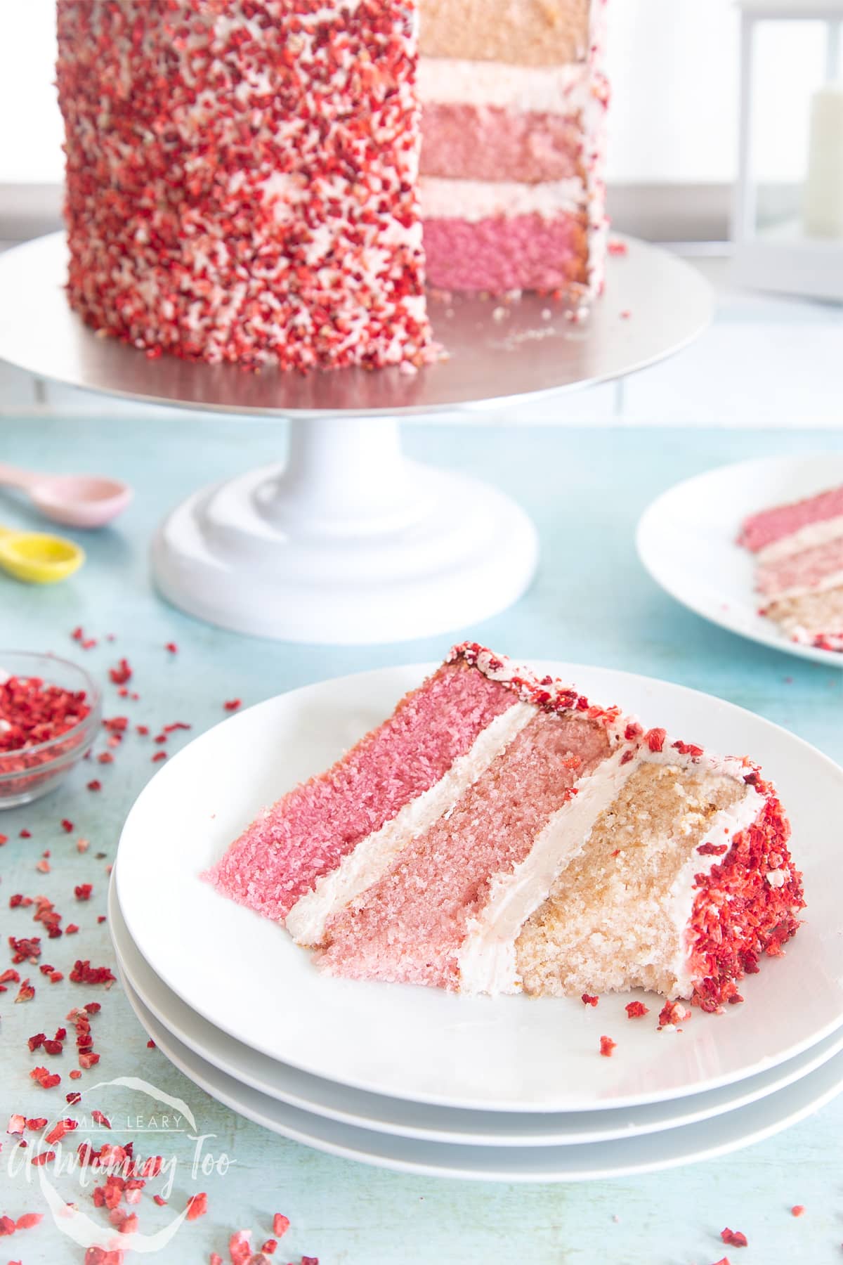 A slice of pink ombre cake on a stack of small white plates. The cake has three layers, each an increasingly intense shade of pink. The sponges are layered with pale pink frosting and the outside is decorated with freeze dried strawberry pieces. A piece of the slice has been cut away.
