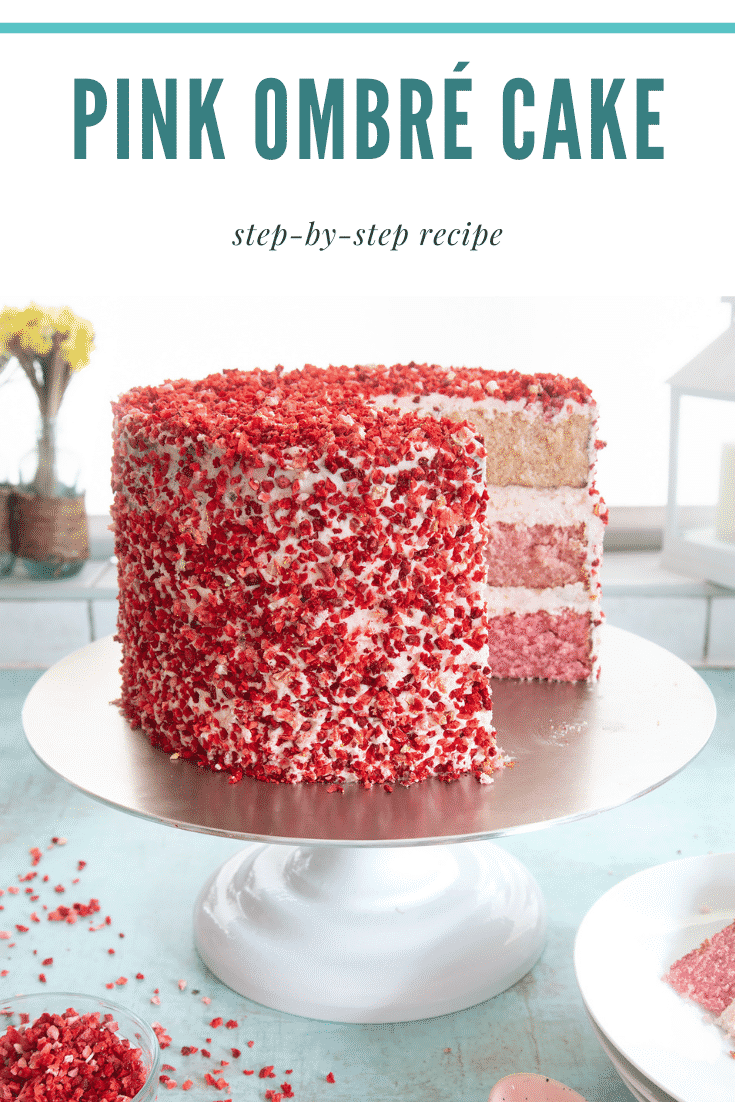 A pink ombre cake on a cake stand. The cake has three layers, each an increasingly intense shade of pink. The sponges are layered with pale pink frosting and the outside is decorated with freeze dried strawberry pieces. Caption reads: pink ombre cake step-by-step recipe