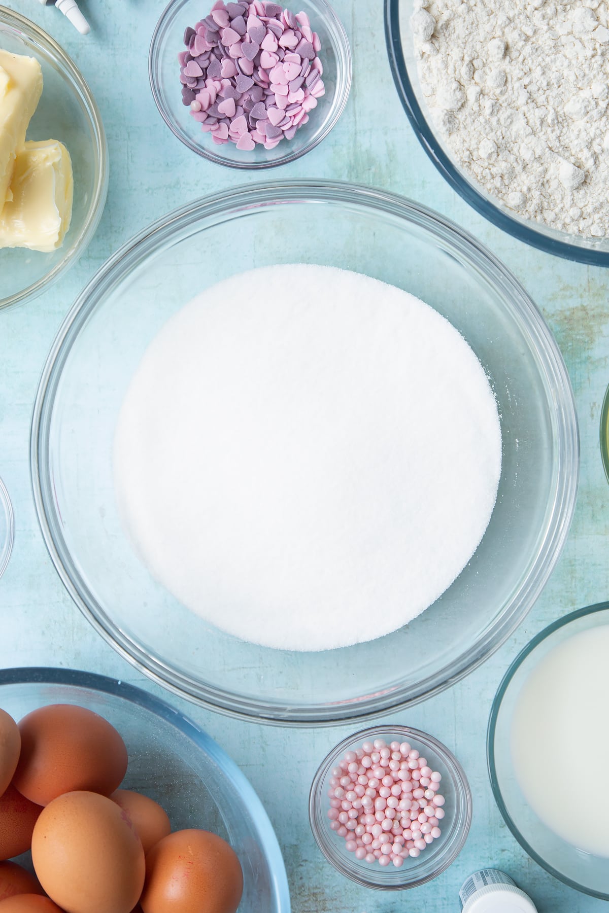 Sugar in a mixing bowl. Ingredients to make pink ombre cake surround the bowl.