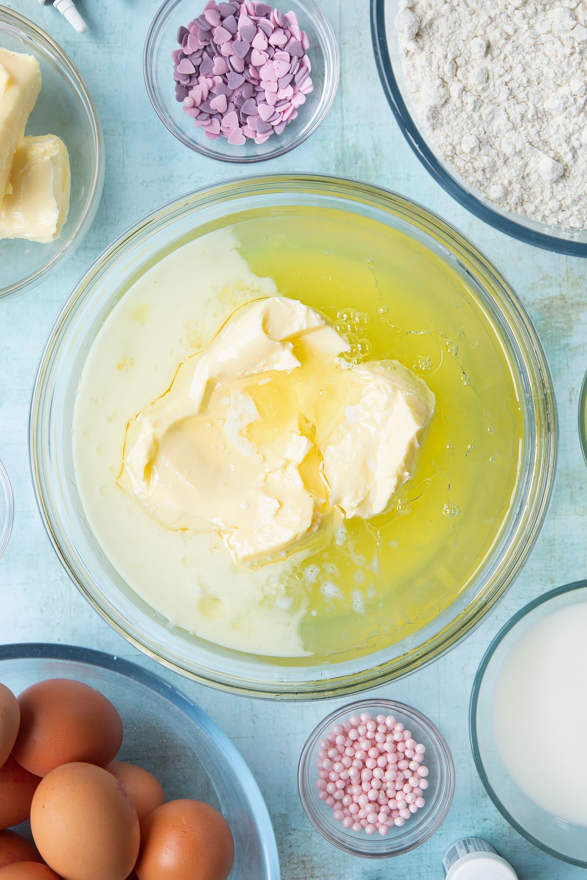 Sugar, egg whites, butter, oil and milk in a mixing bowl. Ingredients to make pink ombre cake surround the bowl.