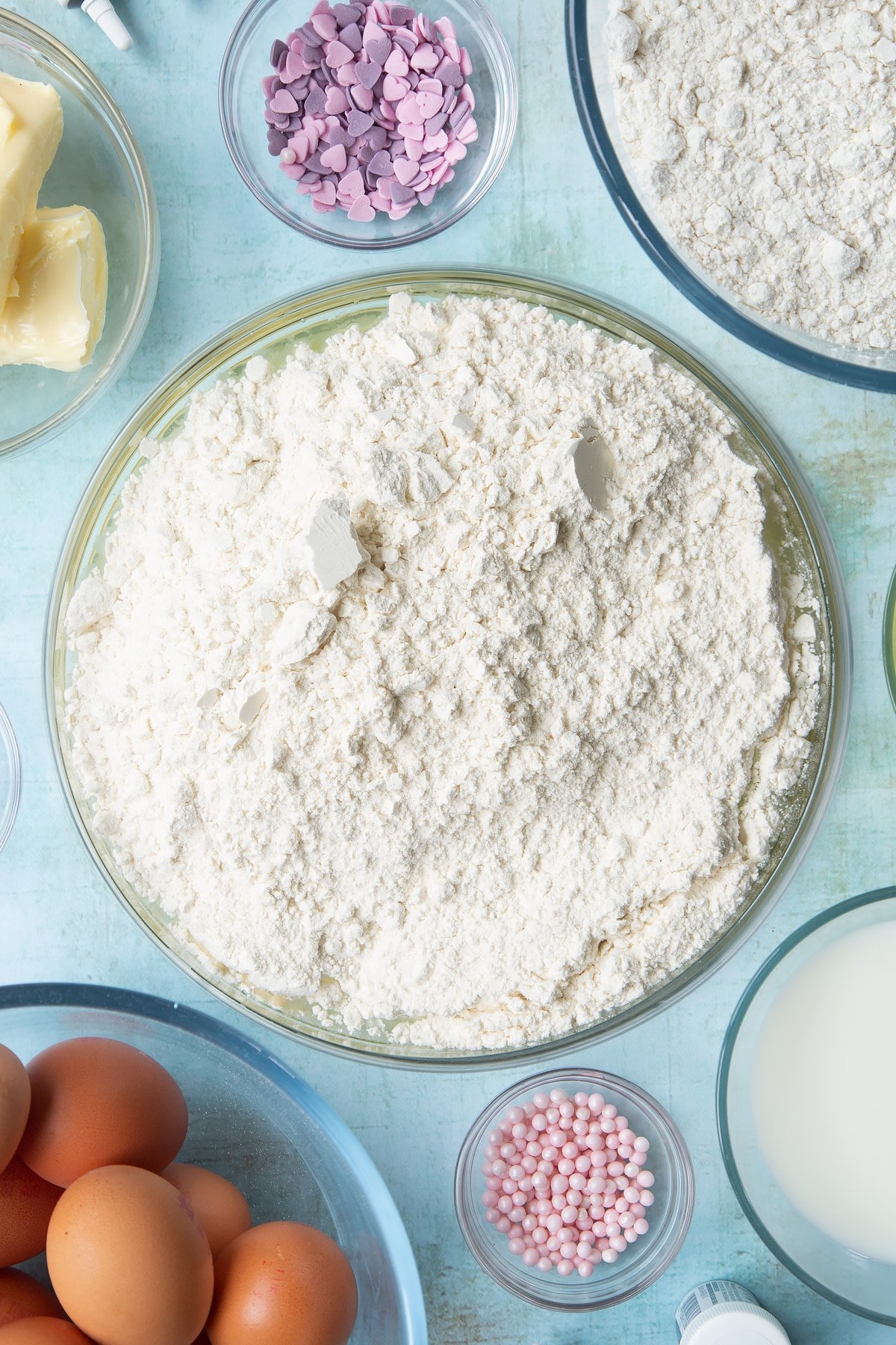 Sugar, egg whites, butter, oil, milk and flour in a mixing bowl. Ingredients to make pink ombre cake surround the bowl.