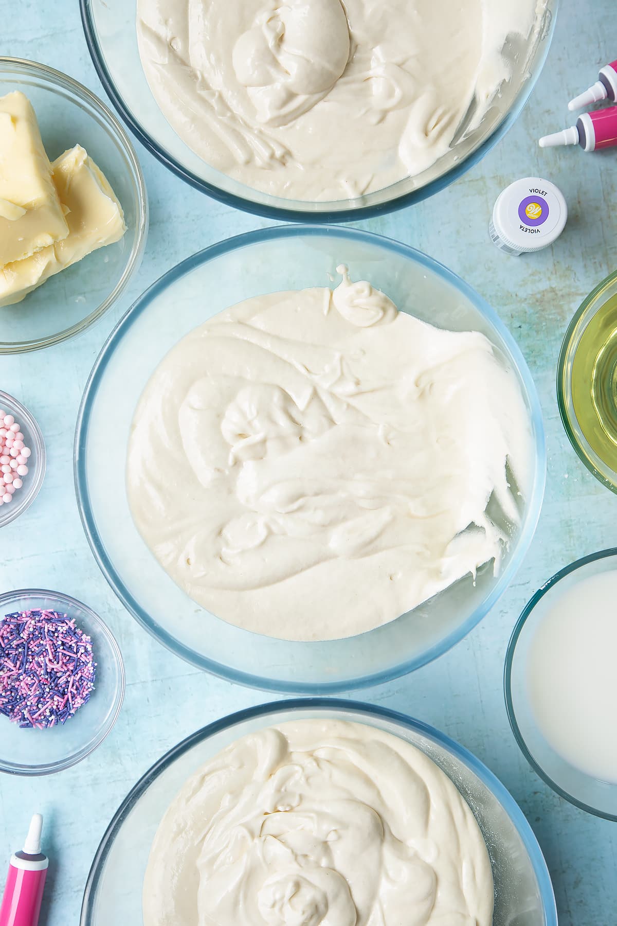 Pale cake batter divided into three mixing bowls. Ingredients to make pink ombre cake surround the bowls.