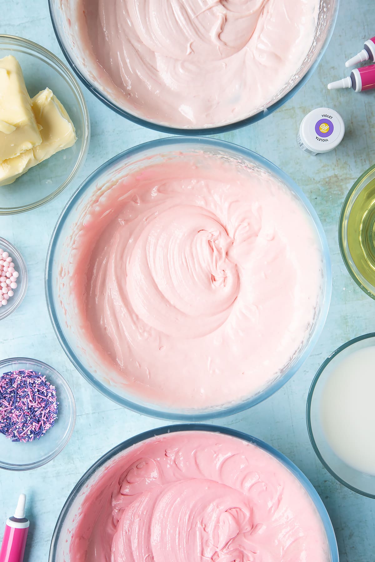 Cake batter divided into three mixing bowls, each dyed a different shade of pink. Ingredients to make pink ombre cake surround the bowls.