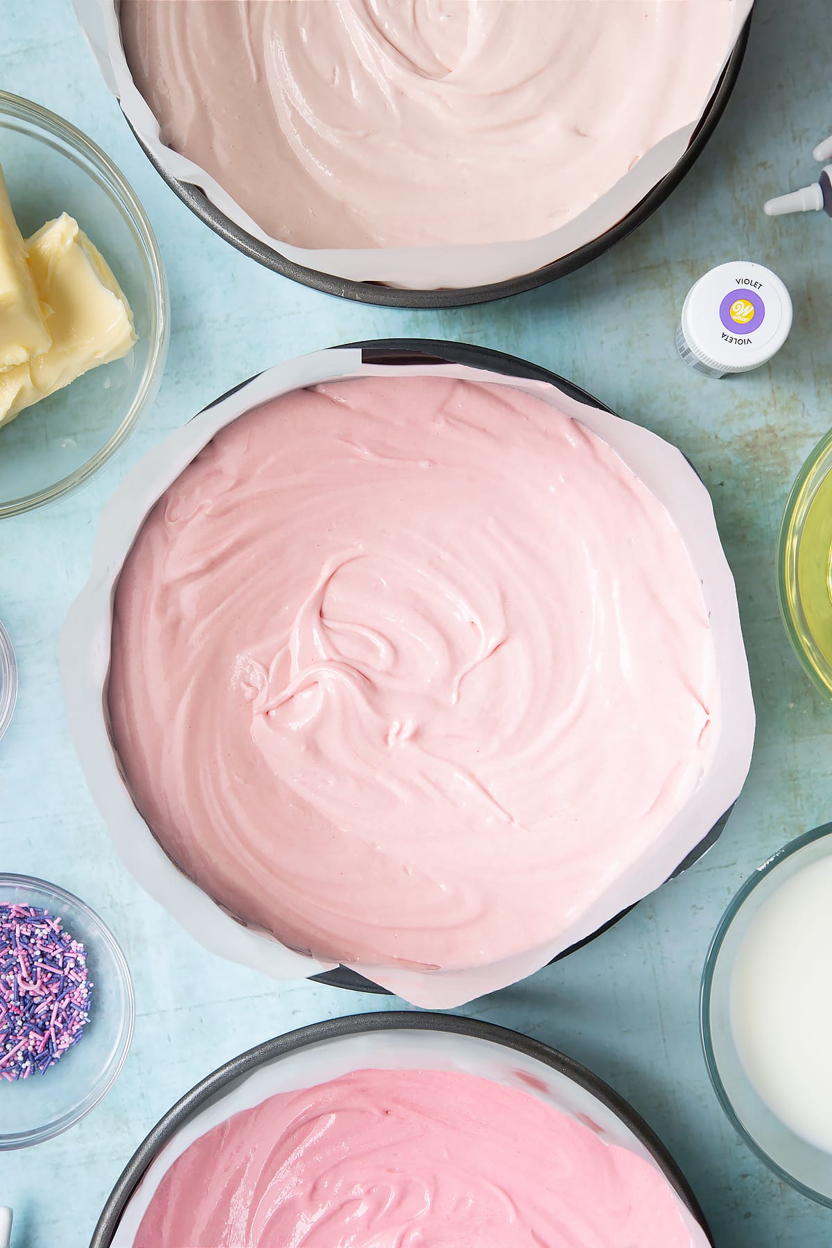 Cake batter divided into three lined cake tins, each dyed a different shade of pink. Ingredients to make pink ombre cake surround the tins.