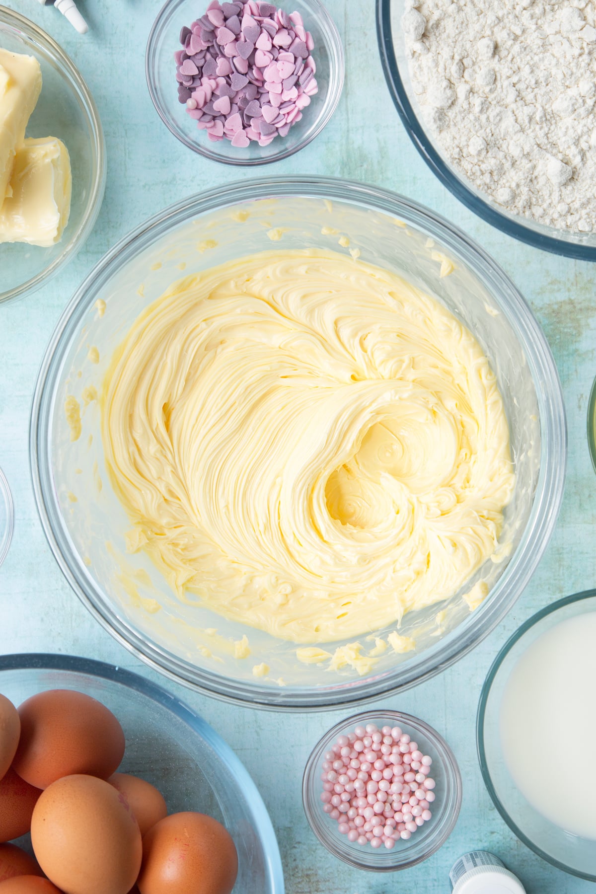 Whipped butter in a mixing bowl. Ingredients to make pink ombre cake surround the bowl.