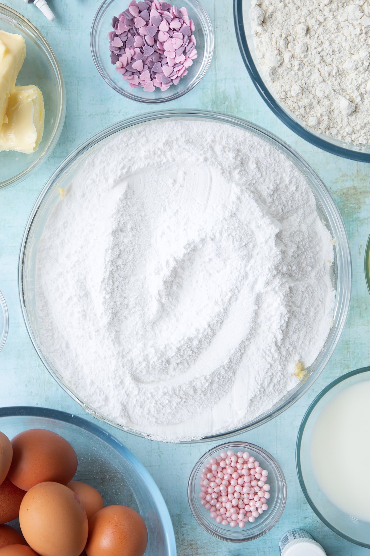 Butter, icing sugar and vanilla in a mixing bowl. Ingredients to make pink ombre cake surround the bowl.