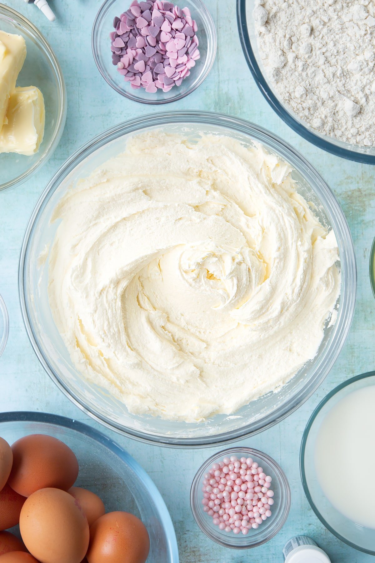 Vanilla buttercream in a mixing bowl. Ingredients to make pink ombre cake surround the bowl.