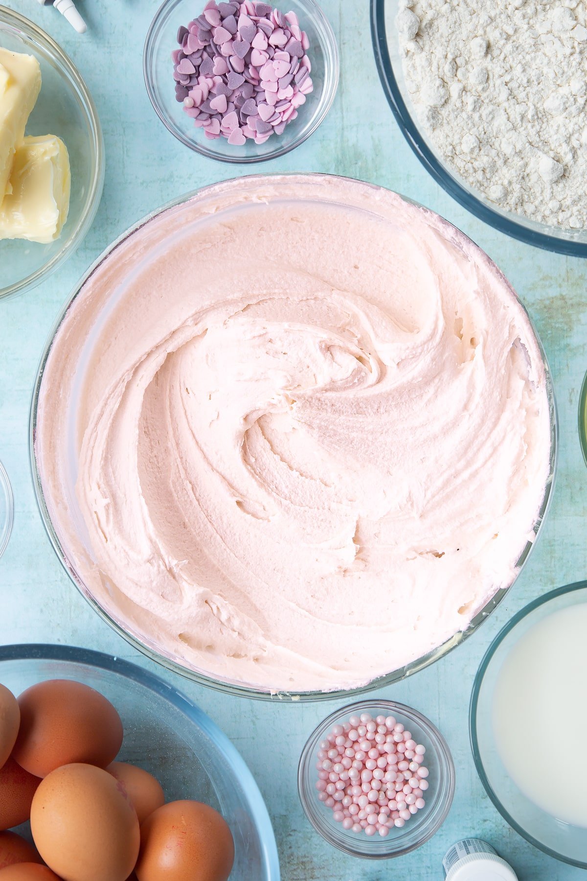 Pale pink vanilla buttercream in a mixing bowl. Ingredients to make pink ombre cake surround the bowl.