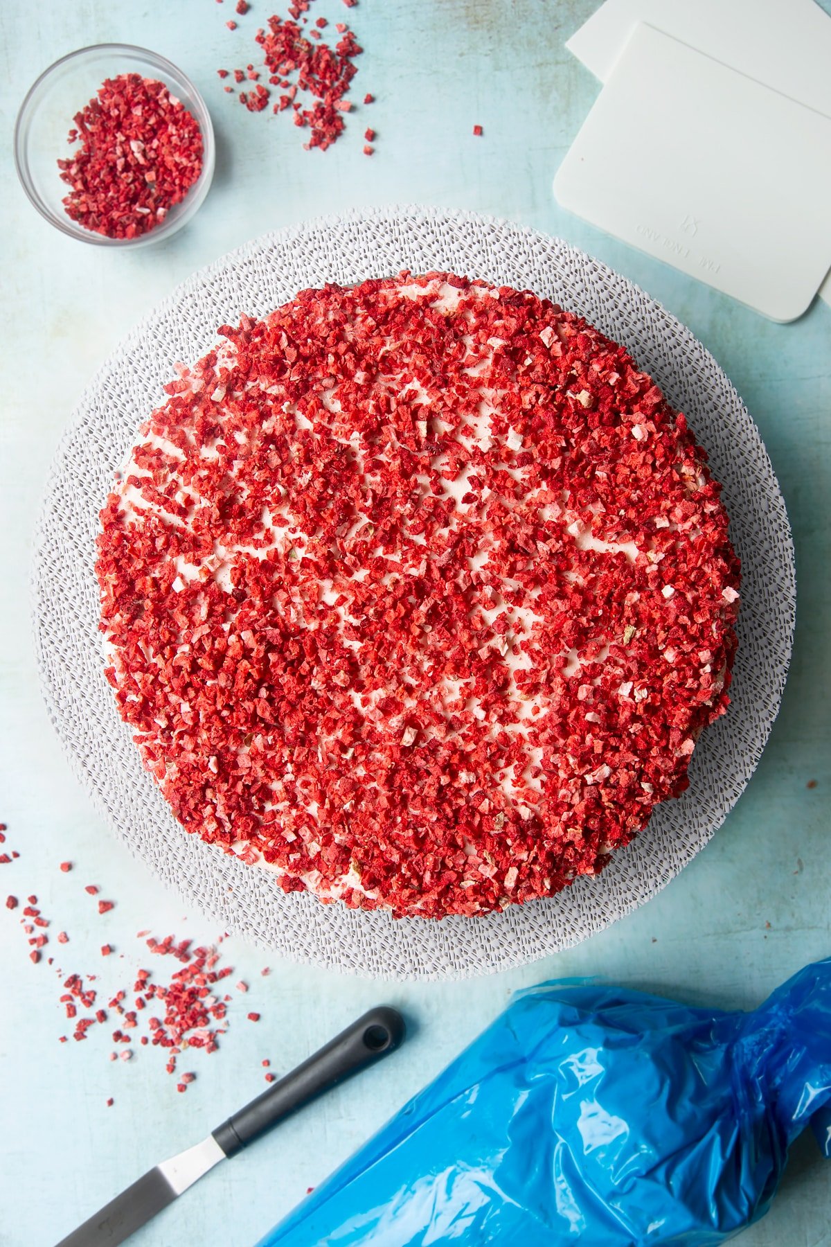 The three layers of a pink ombre cake from brightest to palest pink, stacked on a cake turntable. Pale pink frosting is sandwiched between the layers and coats the outside of the cake, which is then scattered with dried strawberry pieces.