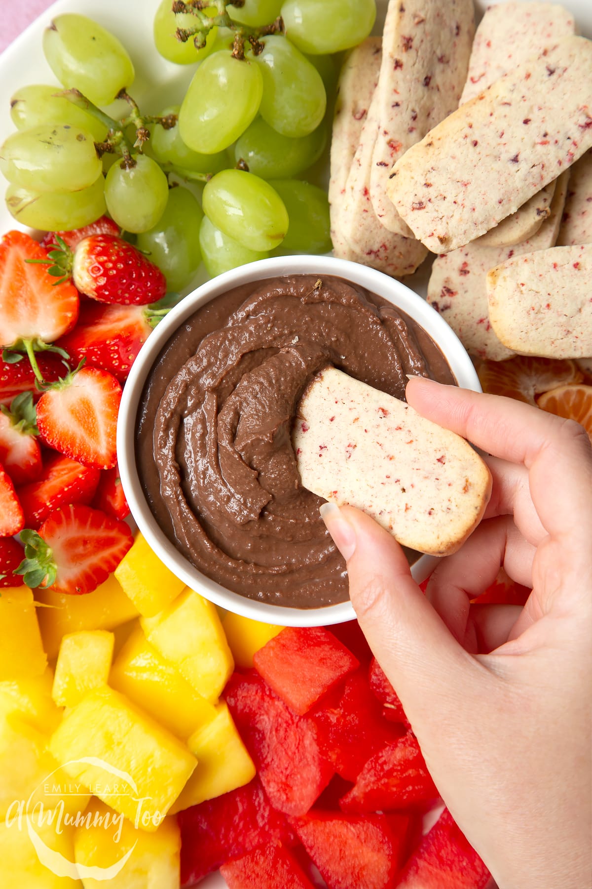 Strawberry biscuits surrounded by grapes, strawberries, mango and watermelon with a chocolate dip in the center. 