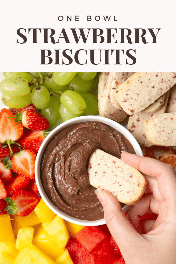 Strawberry biscuit being dipped into a bowl of chocolate spread. In the background there's a range of different fruits and a pile of strawberry biscuits. 