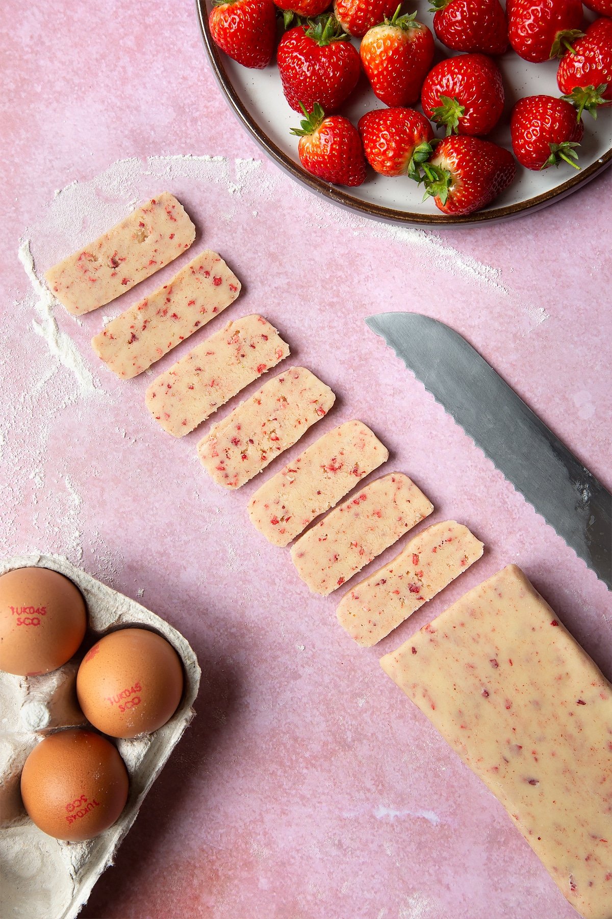 The strawberry biscuit dough cut into slices around 1/4 inch thick. At the side there's a plate of strawberries and a packet of eggs. 