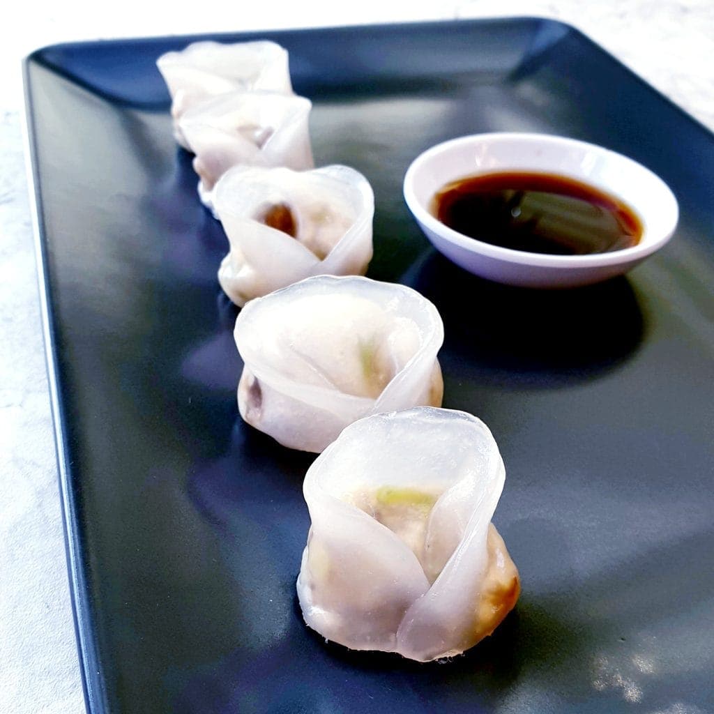 Five tofu dumplings in a line on a black plate with a dipping dish of soy sauce at the side.