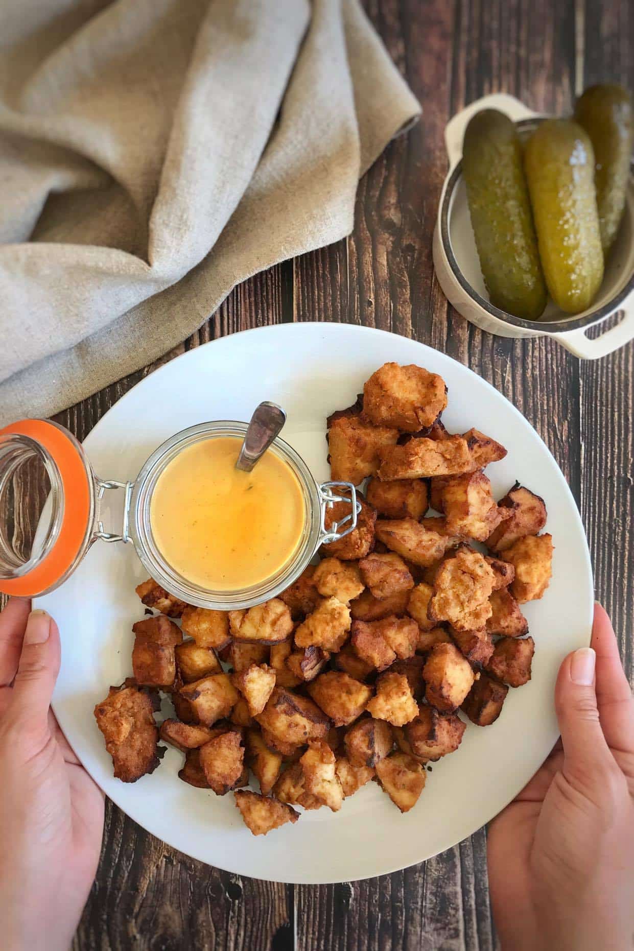Vegan nuggets sit on a plate next to a dish with a dipping sauce on a wooden background. Hands come in to pick up the plate.