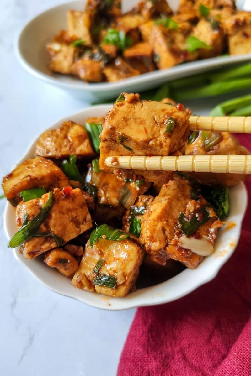 Chilli garlic tofu cubes being picked out of a bowl with a pair of chopsticks.