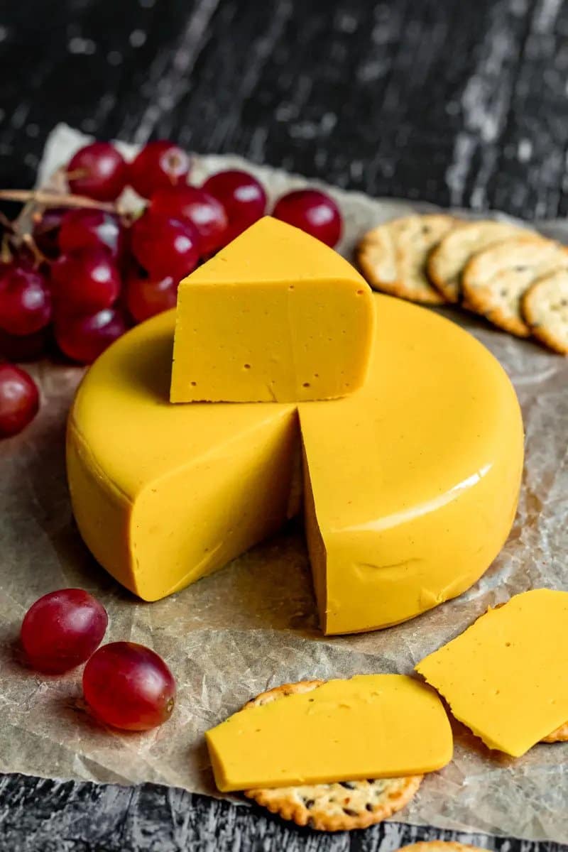 A round block of vegan cheddar cheese which has had a slice cut out. Grapes and a slice of cheese on a cracker sit in the foreground.