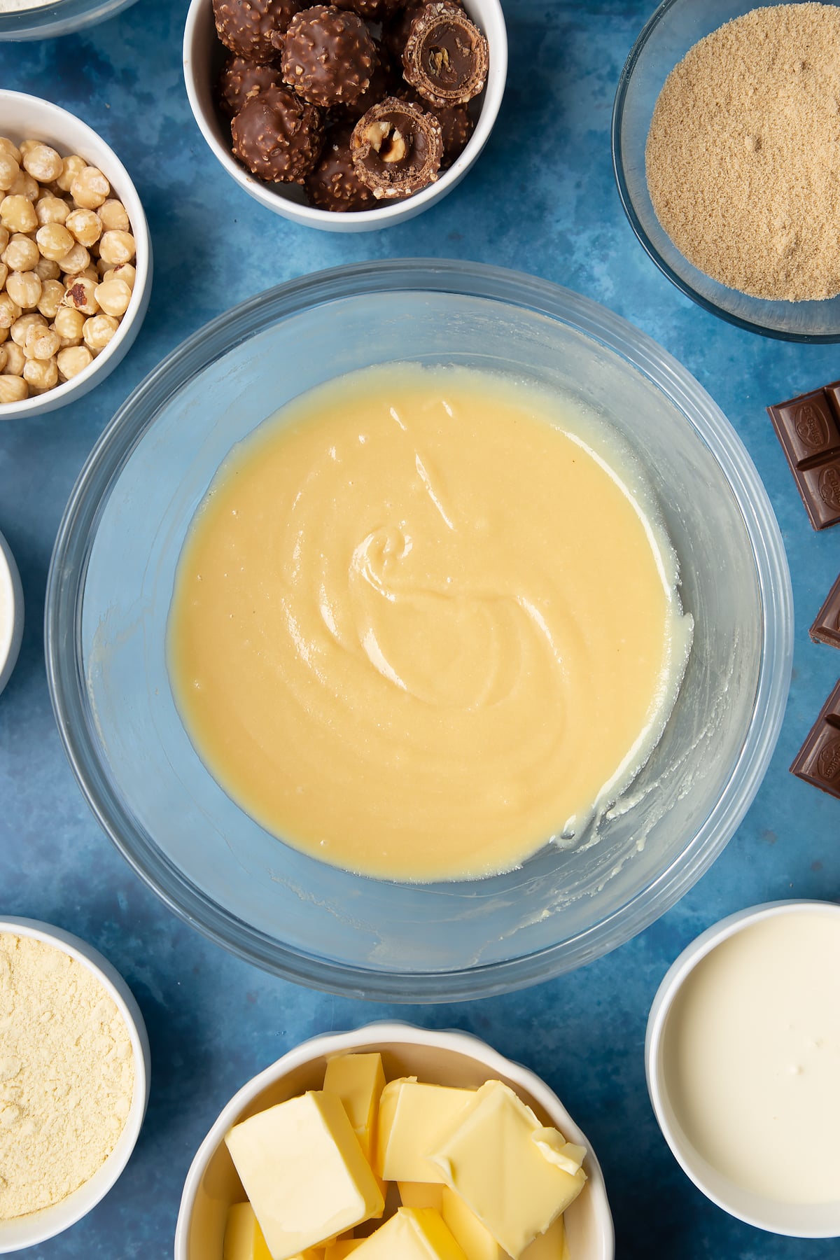 Smooth caramel in a glass mixing bowl. Ingredients to make a Ferrero Rocher slice surround the bowl.