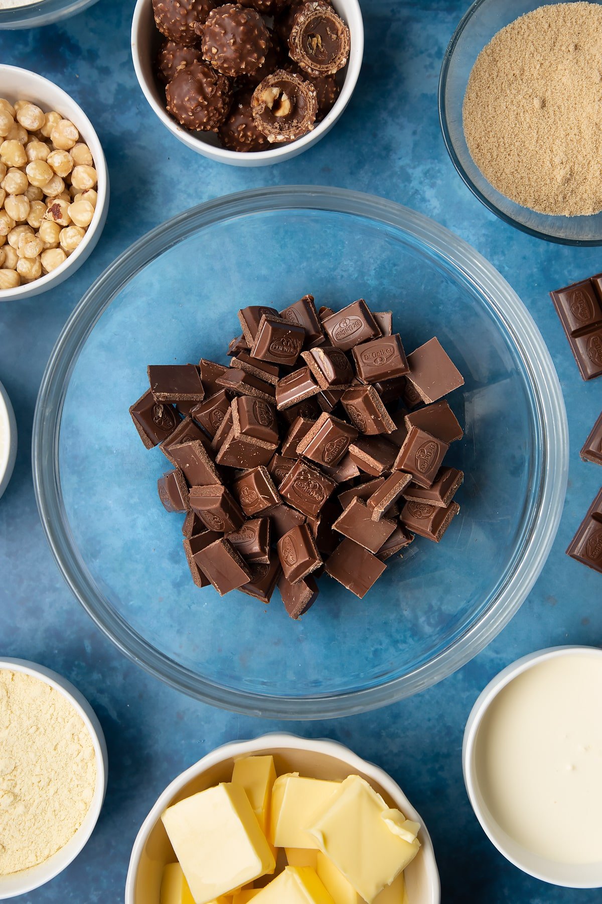 Dark chocolate in a glass mixing bowl. Ingredients to make a Ferrero Rocher slice surround the bowl.