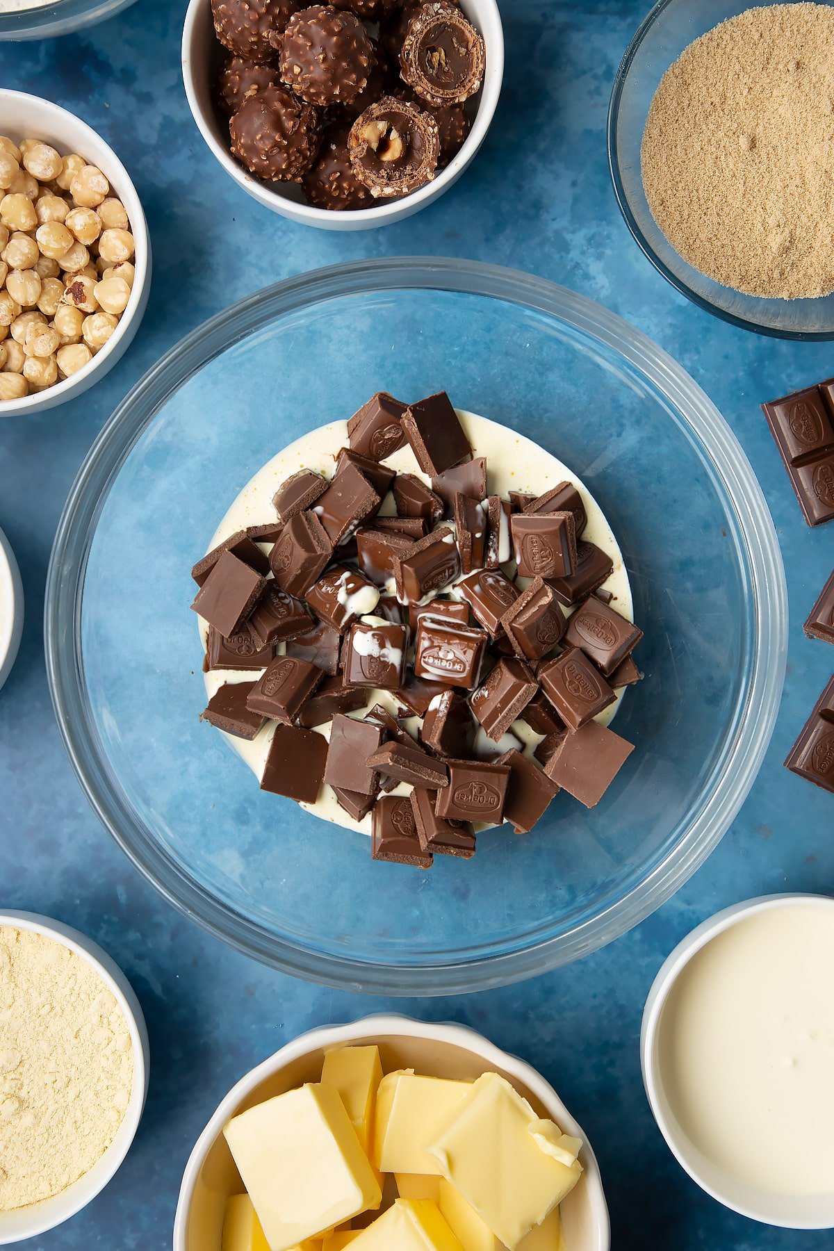 Dark chocolate and hot double cream in a glass mixing bowl. Ingredients to make a Ferrero Rocher slice surround the bowl.