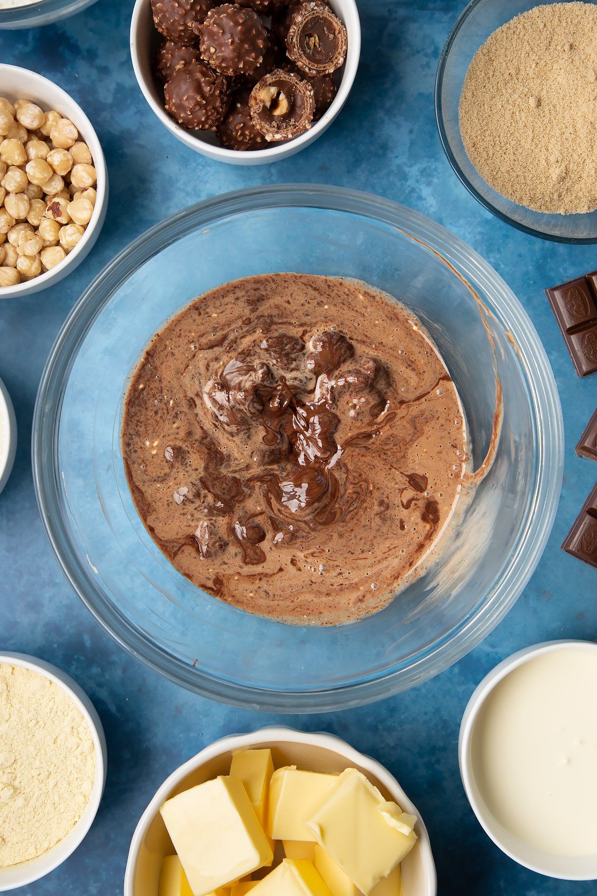 Dark chocolate and hot double cream swirled together in a glass mixing bowl. Ingredients to make a Ferrero Rocher slice surround the bowl.