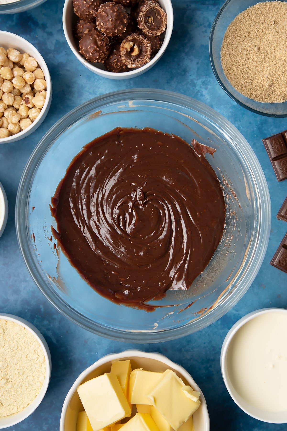 Dark chocolate ganache in a glass mixing bowl. Ingredients to make a Ferrero Rocher slice surround the bowl.