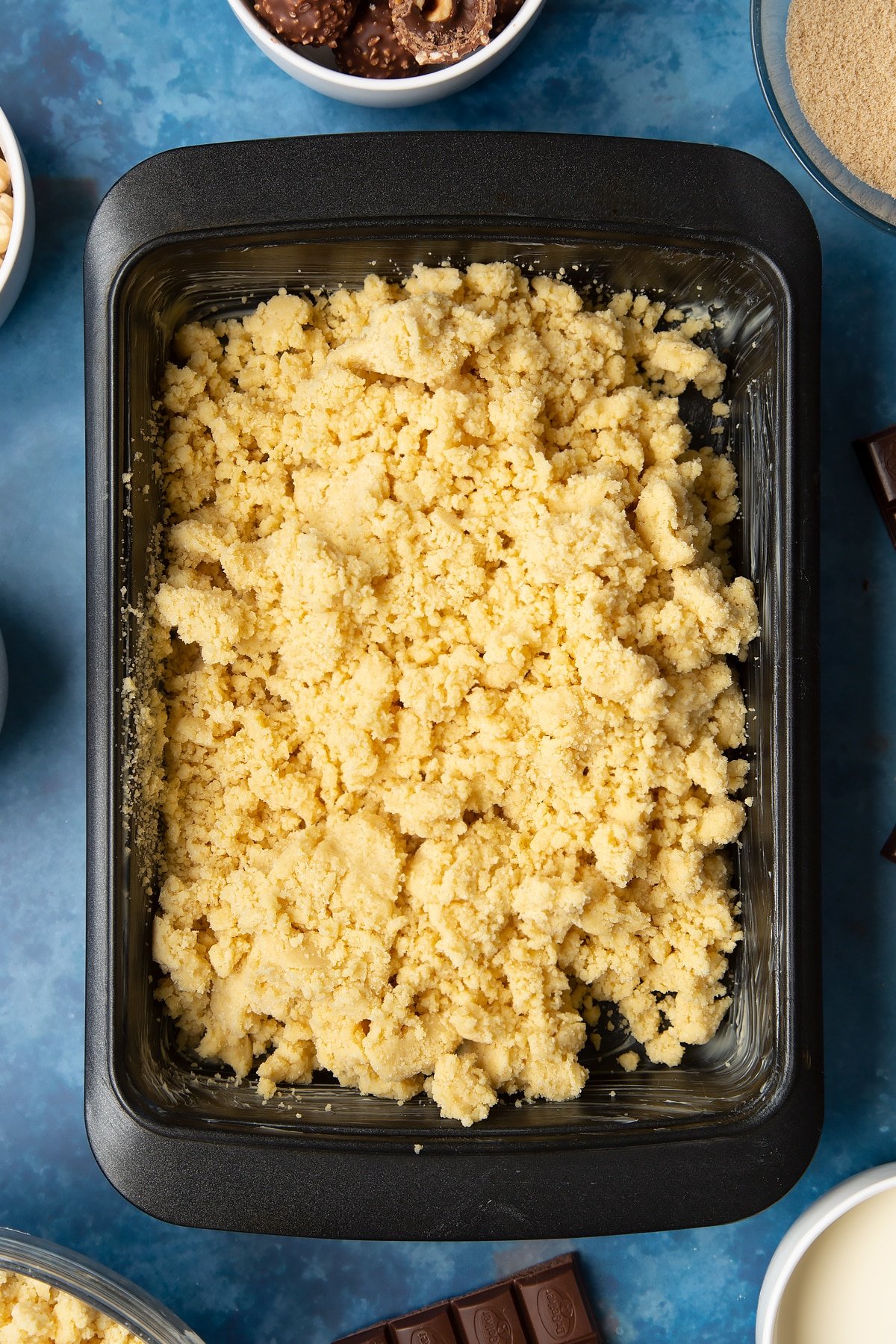 Loose shortbread dough in a greased tray. Ingredients to make a Ferrero Rocher slice surround the tray.