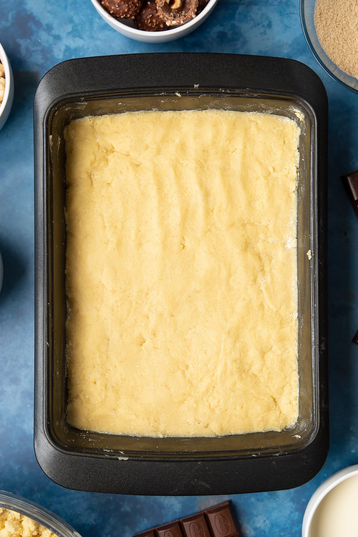 Shortbread dough pressed into a greased tray. Ingredients to make a Ferrero Rocher slice surround the tray.