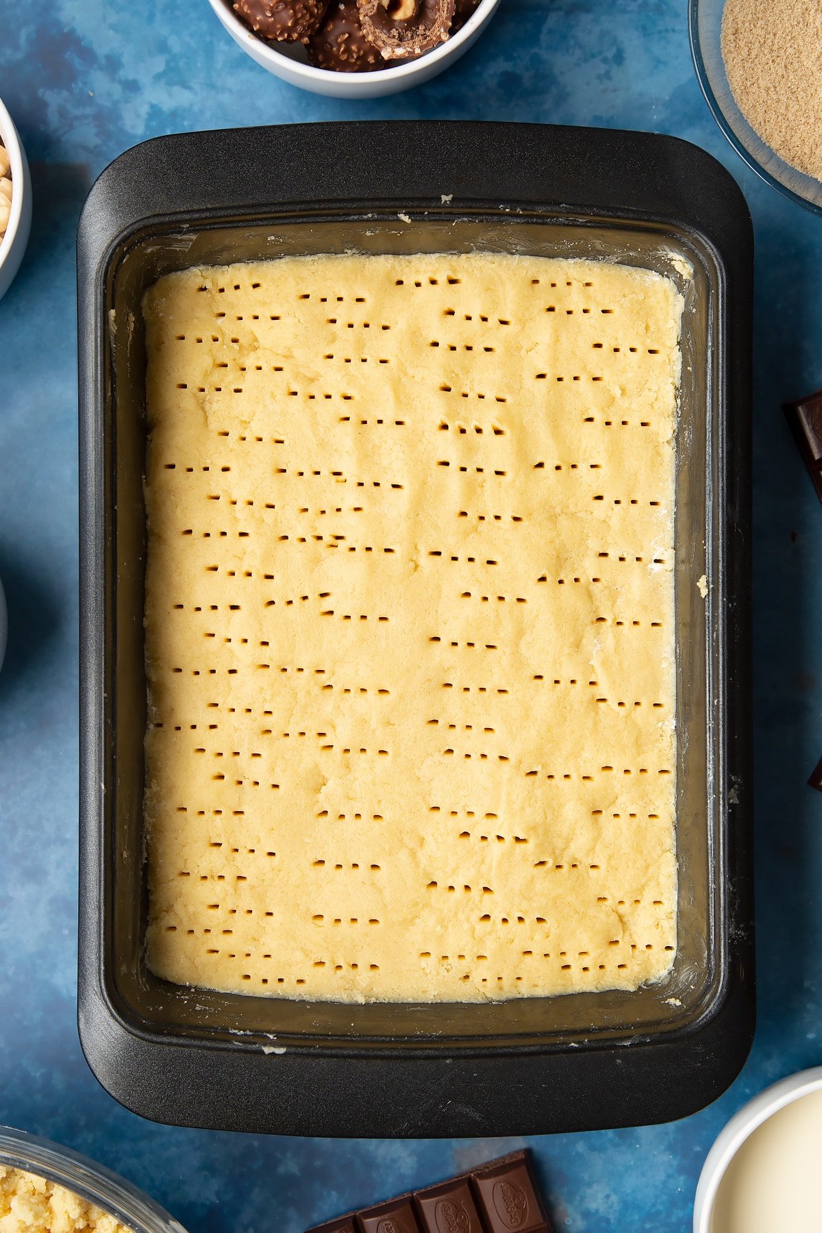 Shortbread dough pressed into a greased tray and pricked with a fork. Ingredients to make a Ferrero Rocher slice surround the tray.