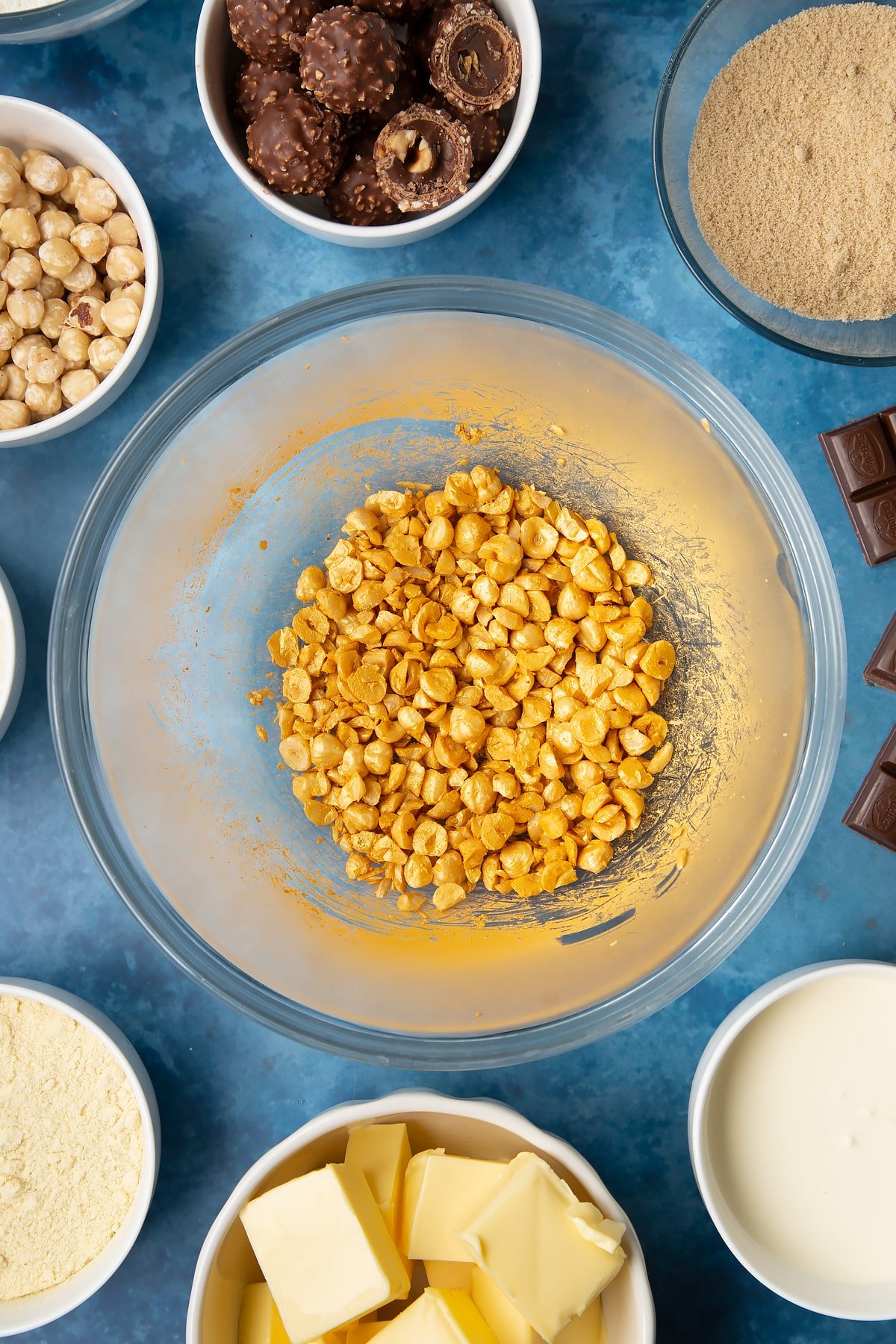 Chopped hazelnuts sprayed gold in a glass mixing bowl. Ingredients to make a Ferrero Rocher slice surround the bowl.