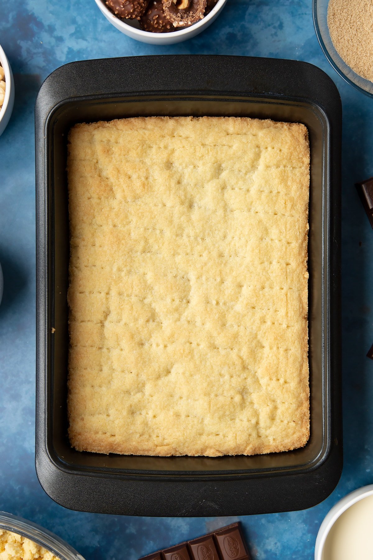 Cooked and cooled shortbread in a tray. Ingredients to make a Ferrero Rocher slice surround the tray.