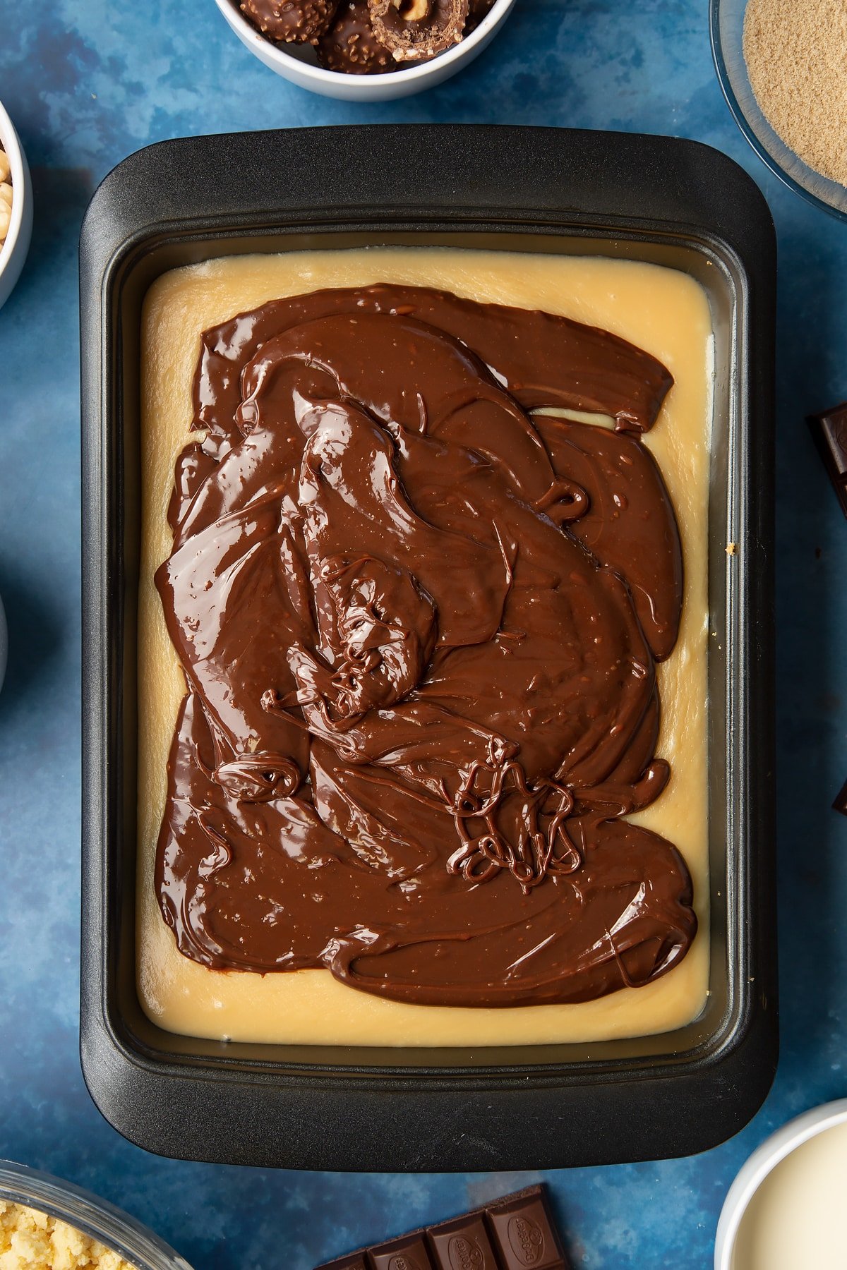 Shortbread topped with caramel in a tray. Dark chocolate ganache is piled on top. Ingredients to make a Ferrero Rocher slice surround the tray.