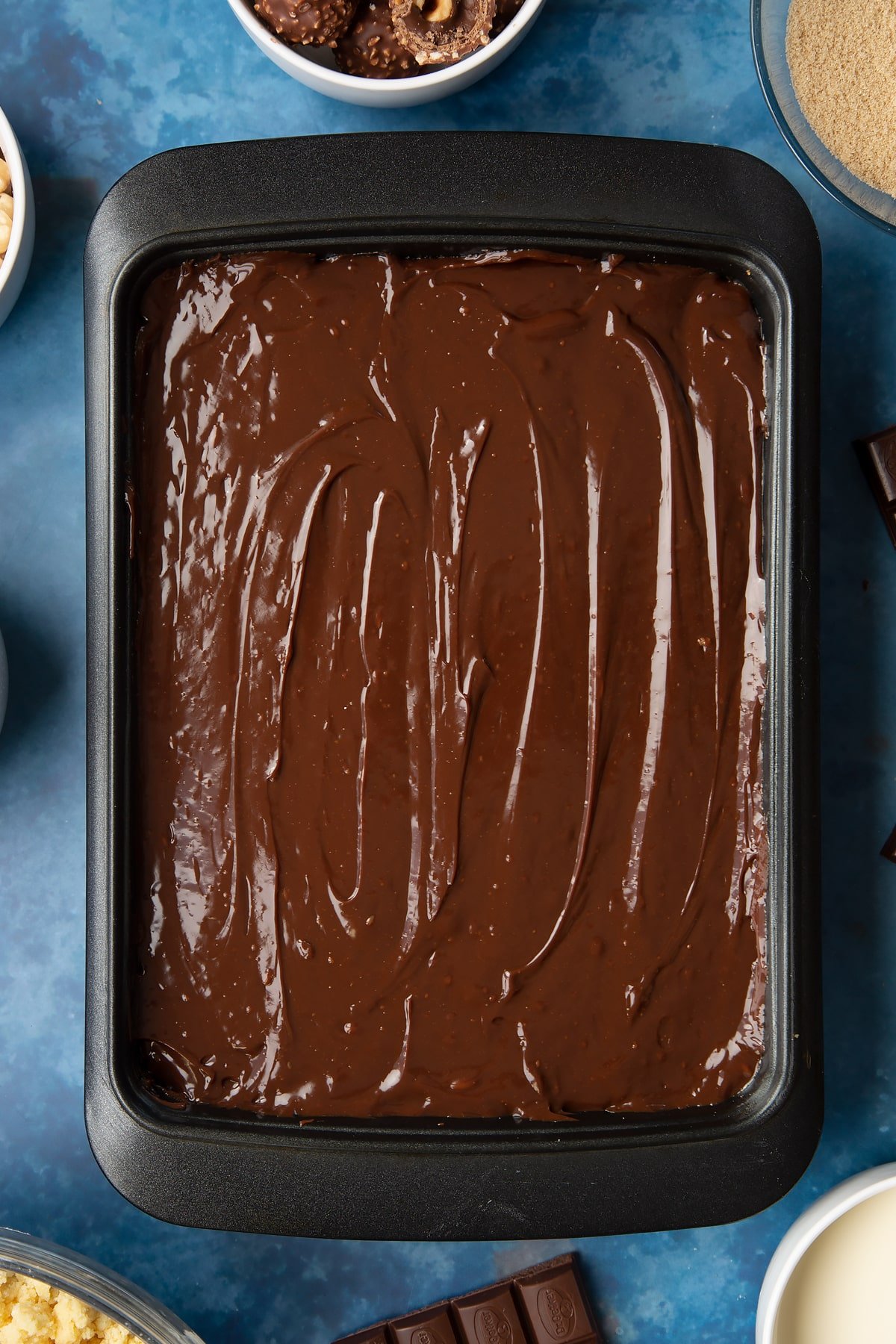 Shortbread topped with caramel and then dark chocolate ganache in a tray. Ingredients to make a Ferrero Rocher slice surround the tray.