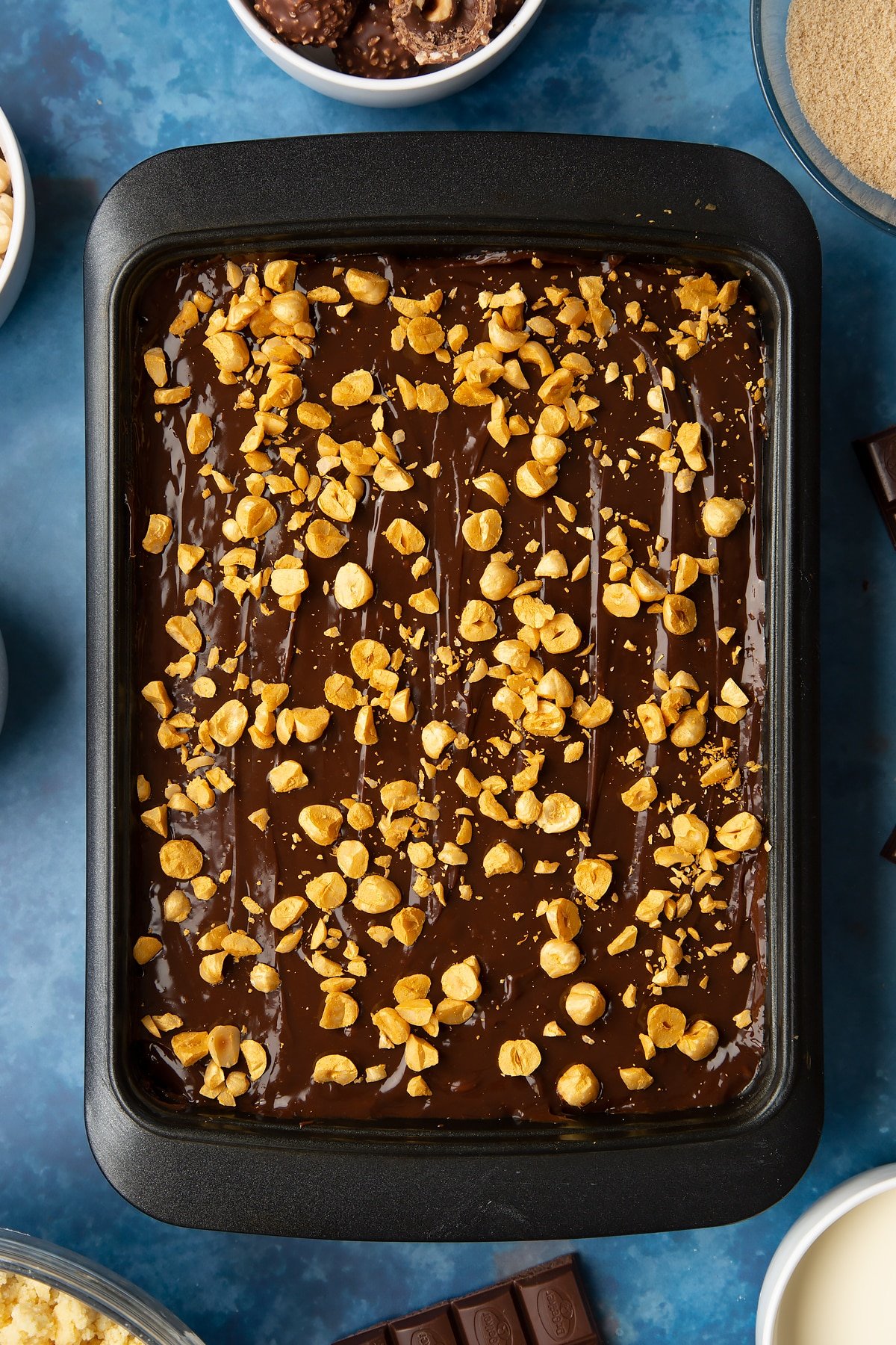 Millionaire's shortbread scatter with golden hazelnuts. Ingredients to make a Ferrero Rocher slice surround the tray.