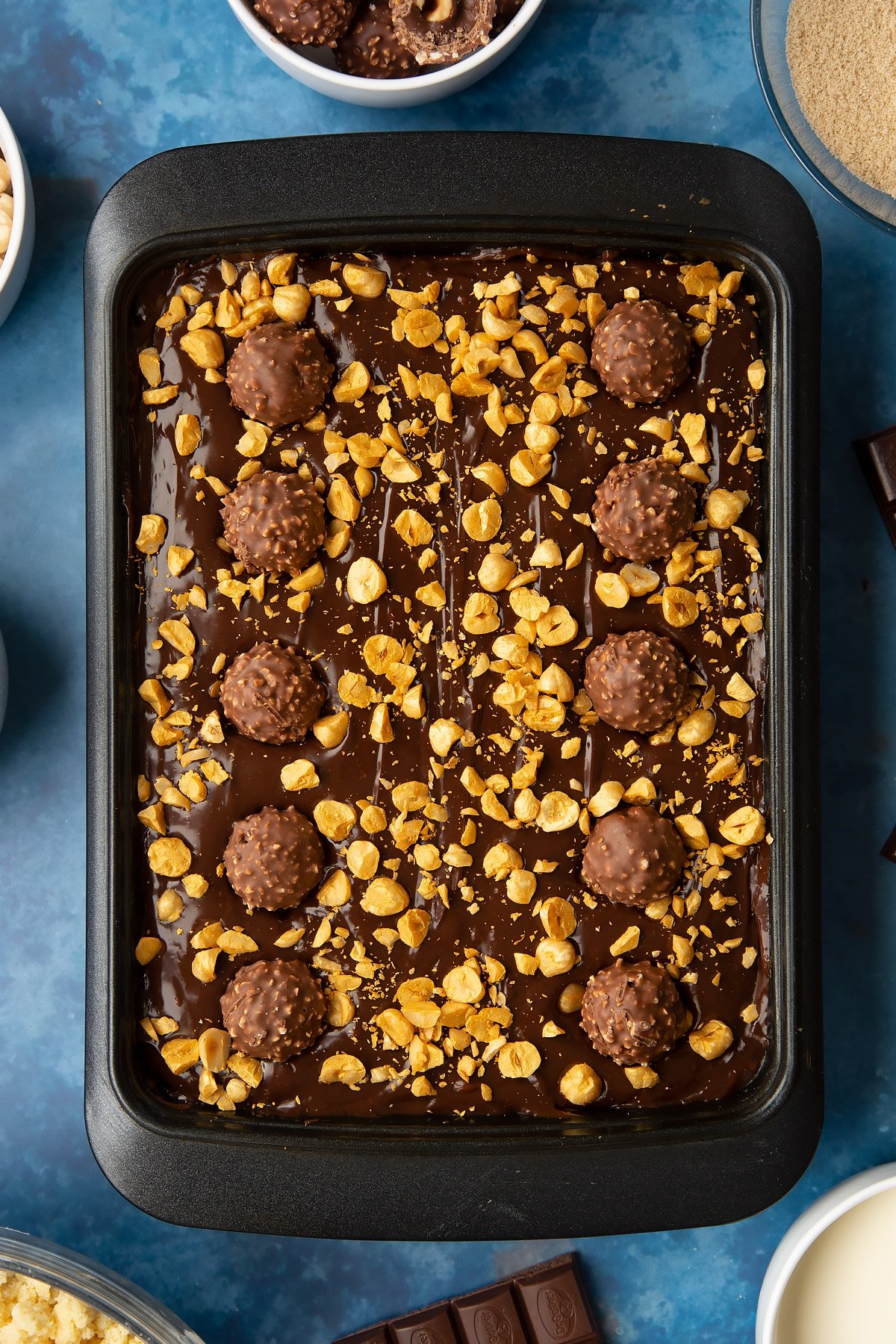 Millionaire's shortbread scatter with golden hazelnuts and topped with Ferrero Rocher. Ingredients to make a Ferrero Rocher slice surround the tray.
