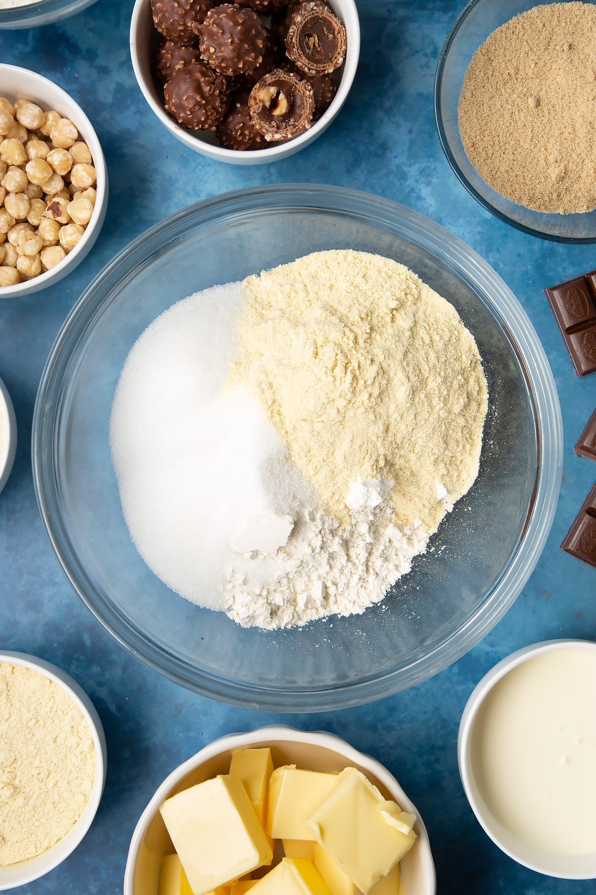 Flour, sugar and semolina in a glass mixing bowl. Ingredients to make a Ferrero Rocher slice surround the bowl.