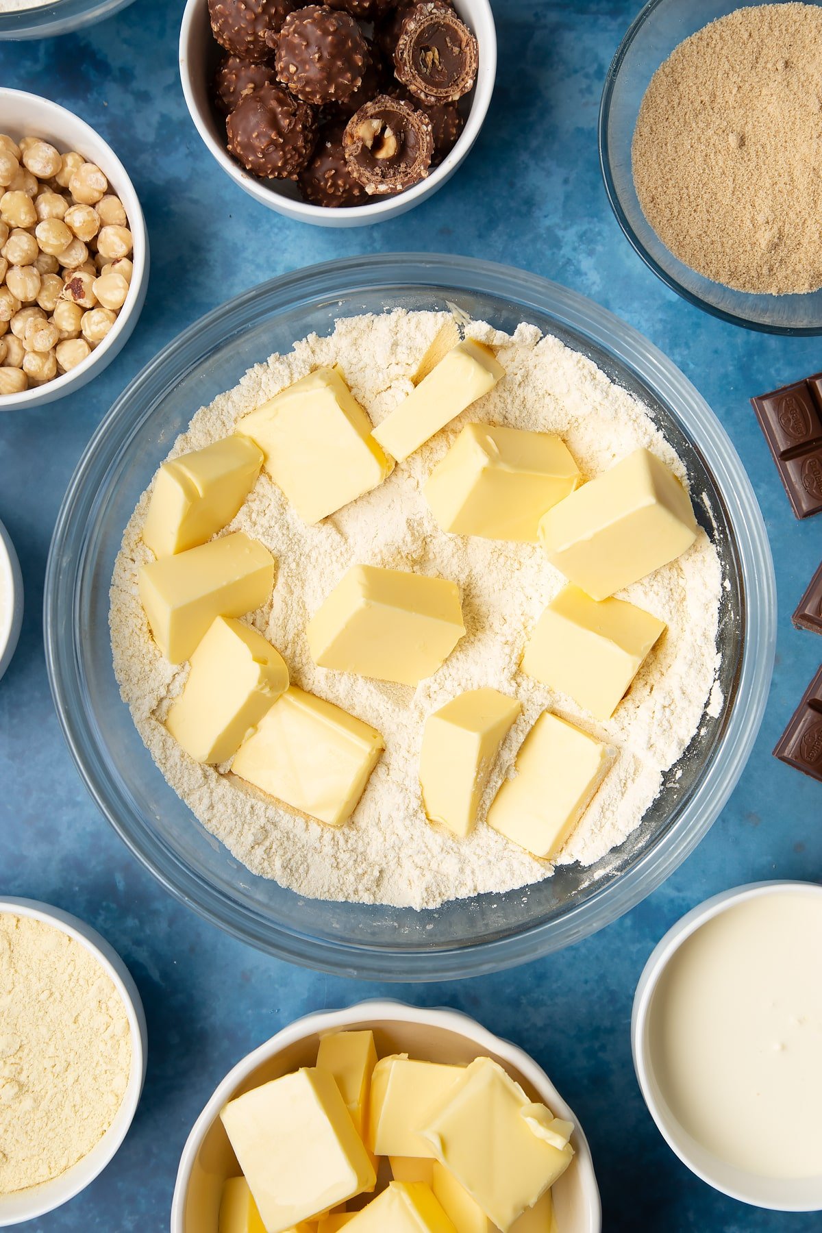 Flour, sugar, semolina and butter in a glass mixing bowl. Ingredients to make a Ferrero Rocher slice surround the bowl.