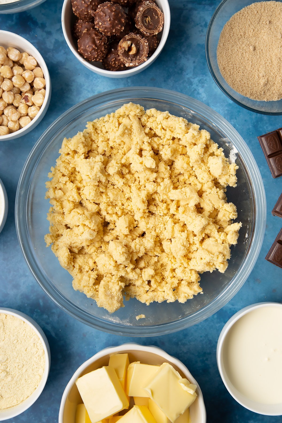 Shortbread dough in a glass mixing bowl. Ingredients to make a Ferrero Rocher slice surround the bowl.