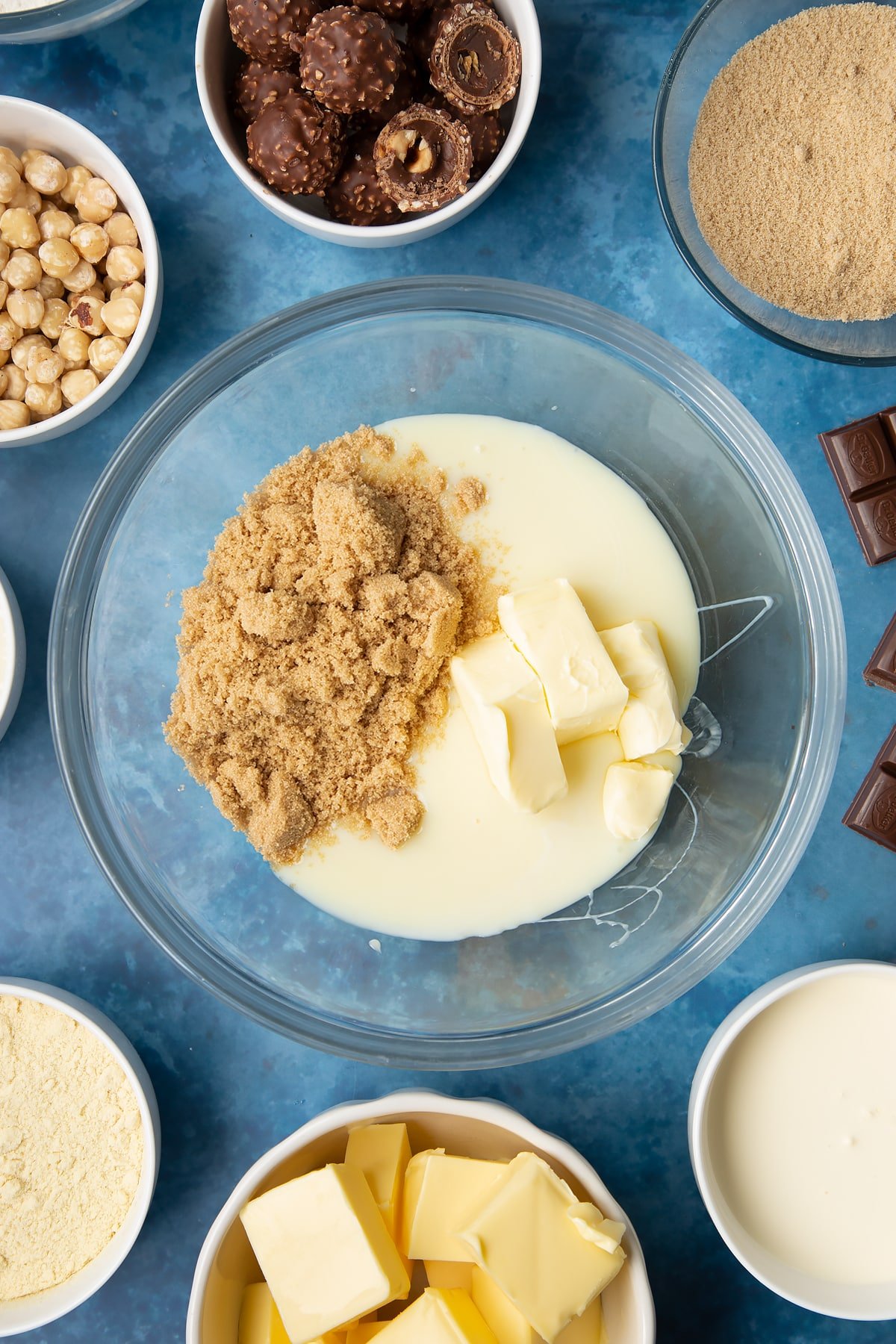 Brown sugar, butter and condensed milk in a glass mixing bowl. Ingredients to make a Ferrero Rocher slice surround the bowl.