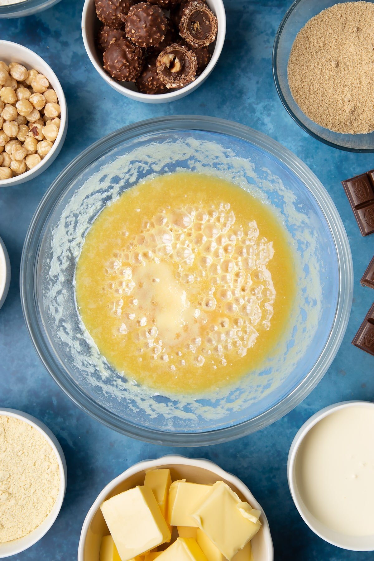 Bubbling caramel in a glass mixing bowl. Ingredients to make a Ferrero Rocher slice surround the bowl.