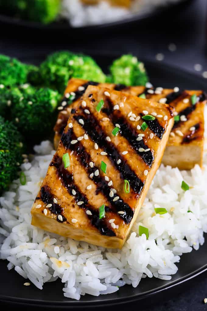 Grilled tofu topped with sesame seeds sits inside a black bowl with broccoli on the side. 