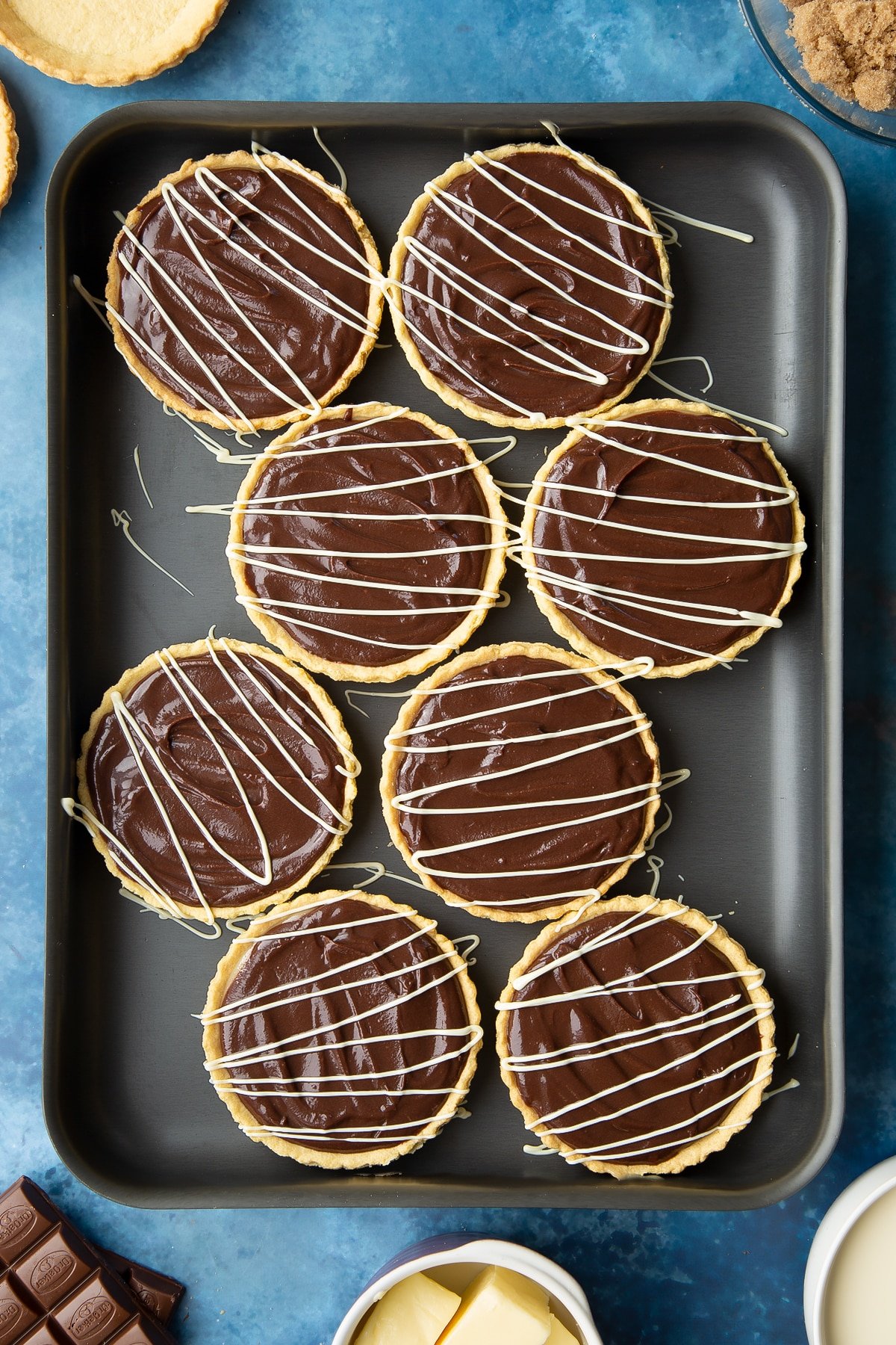 Mini chocolate tarts decorated with drizzled white chocolate on a large metal tray. 