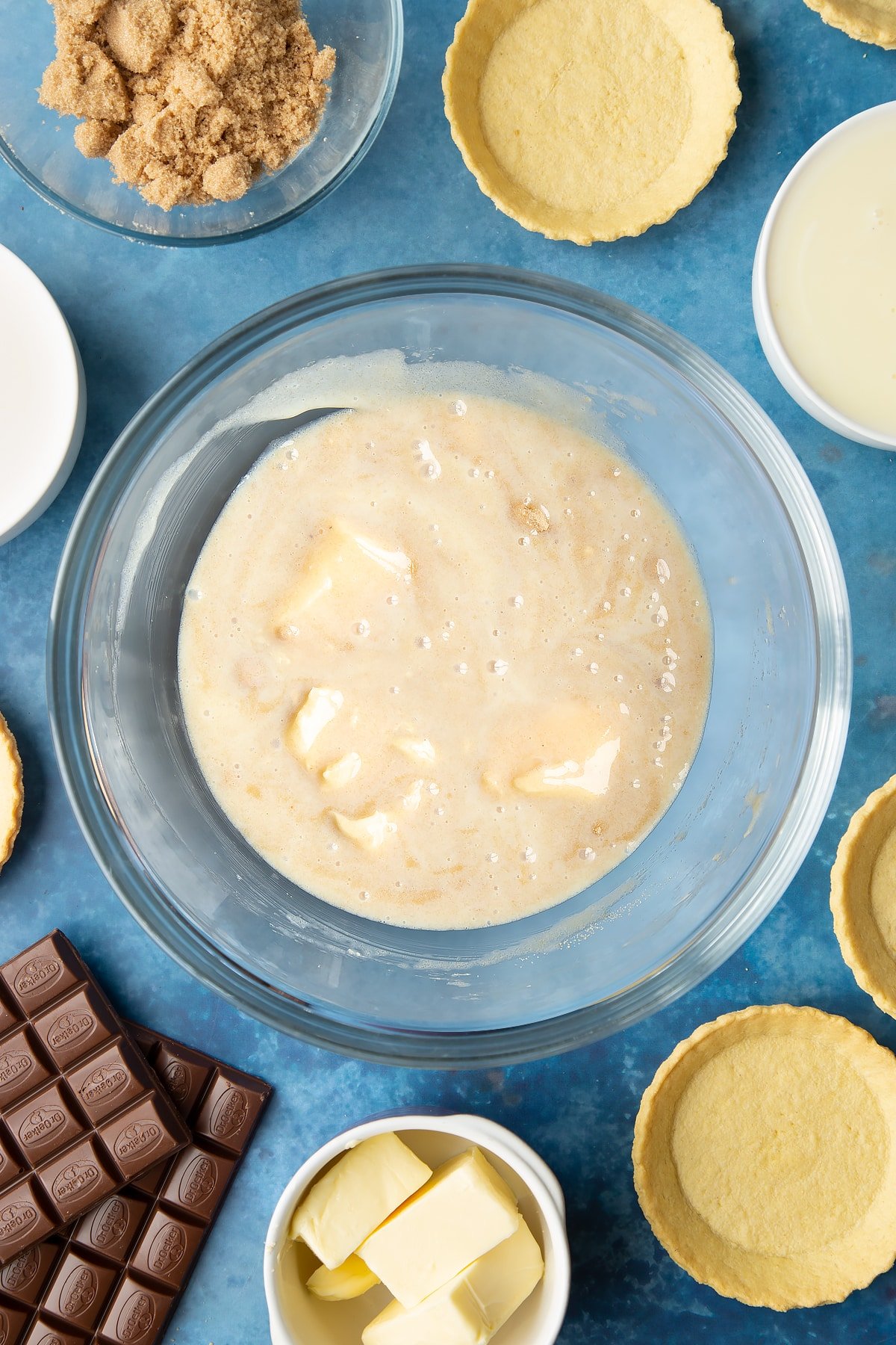 Light brown sugar, butter and condensed milk stirred together in a large mixing bowl. Ingredients to make mini chocolate tarts surround the bowl. 