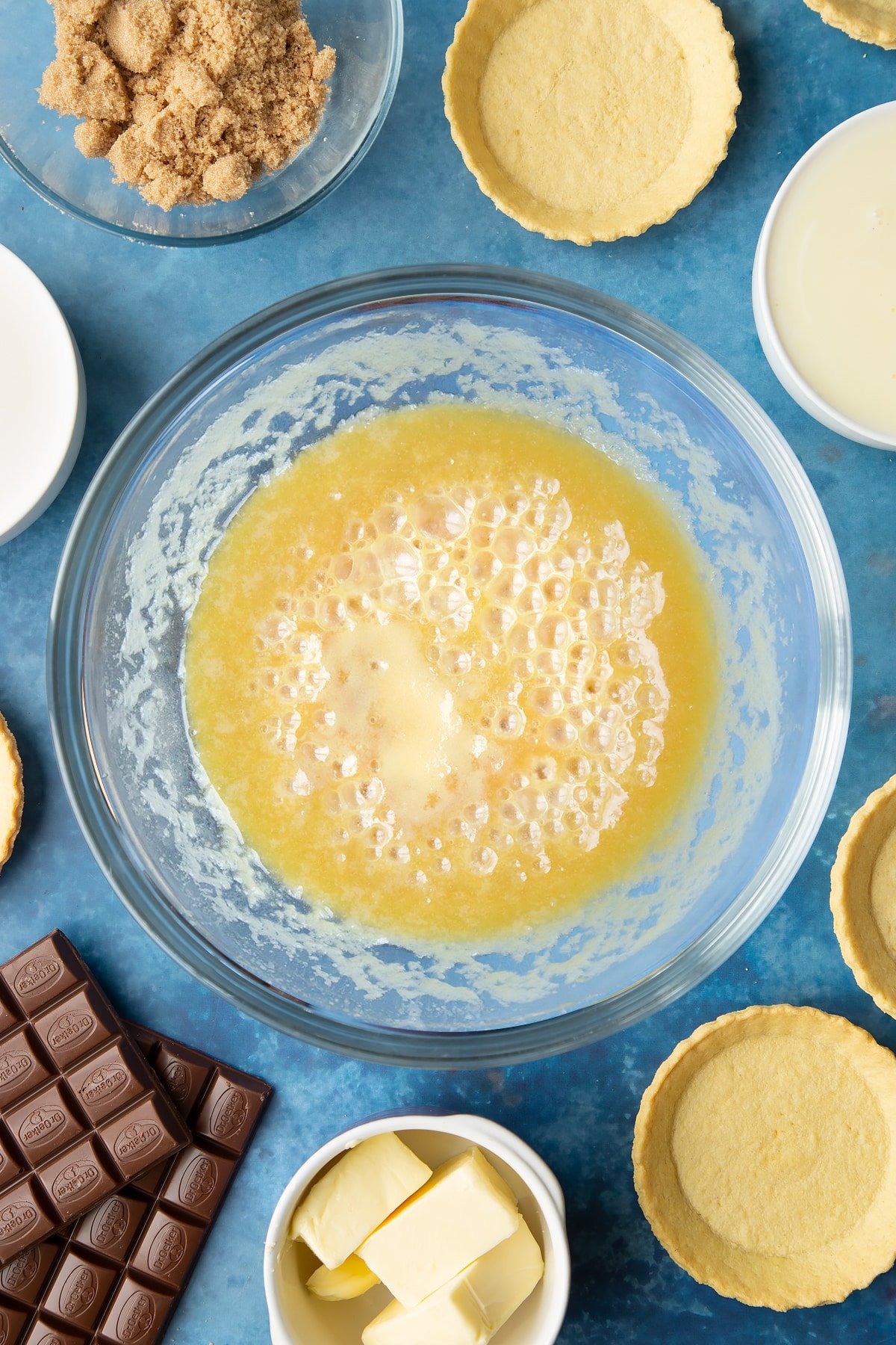 Light brown sugar, butter and condensed milk heated together in a large mixing bowl. Ingredients to make mini chocolate tarts surround the bowl. 