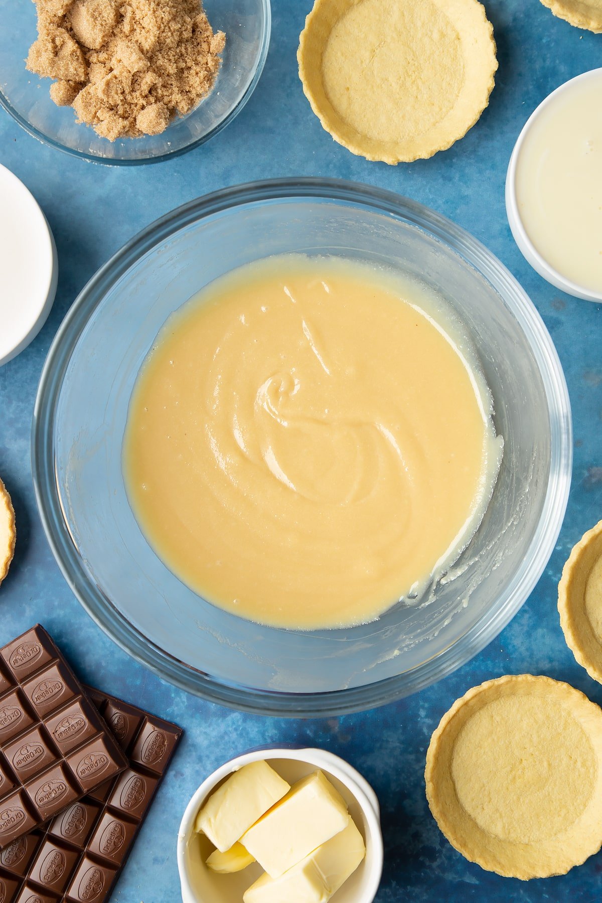 Freshly made caramel in a large mixing bowl. Ingredients to make mini chocolate tarts surround the bowl. 