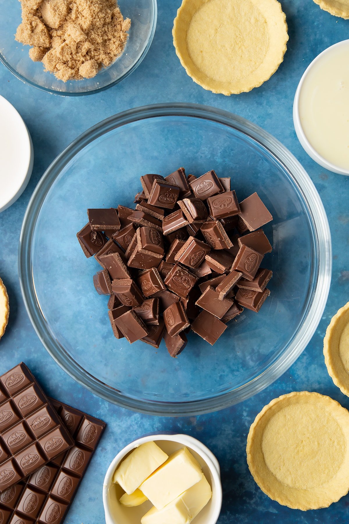 Broken up milk chocolate in a large mixing bowl. Ingredients to make mini chocolate tarts surround the bowl. 