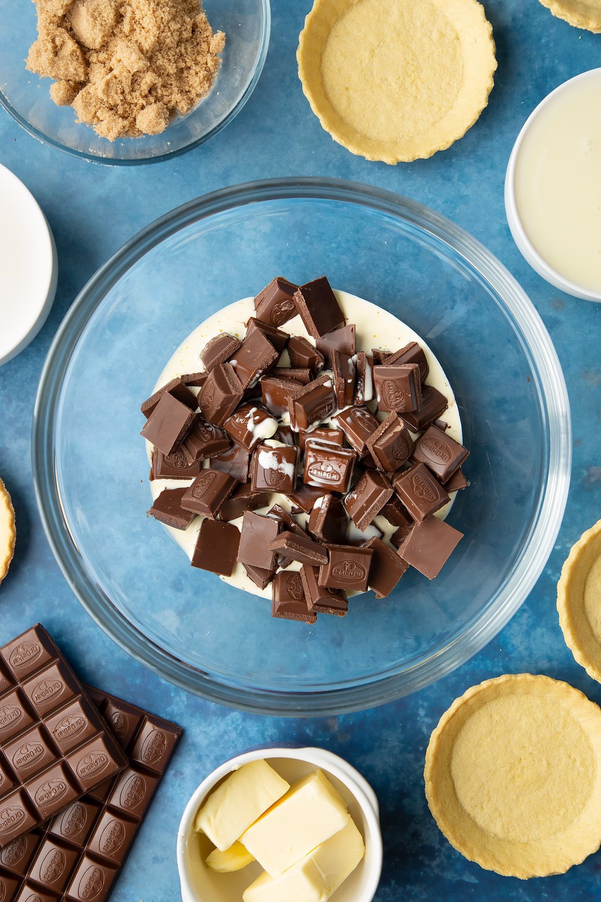 Broken up milk chocolate and heated cream in a large mixing bowl. Ingredients to make mini chocolate tarts surround the bowl. 