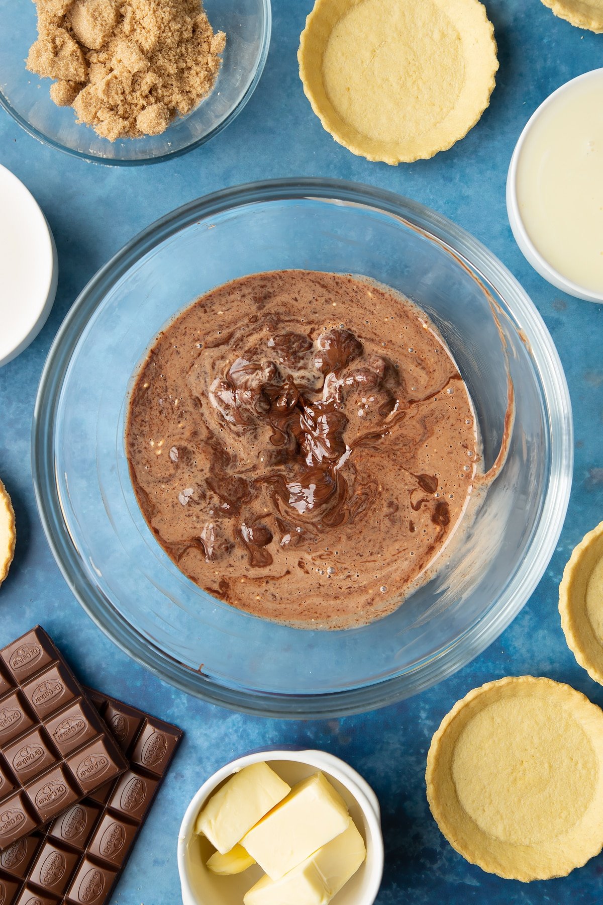 Broken up milk chocolate and heated cream swirled together in a large mixing bowl. Ingredients to make mini chocolate tarts surround the bowl. 