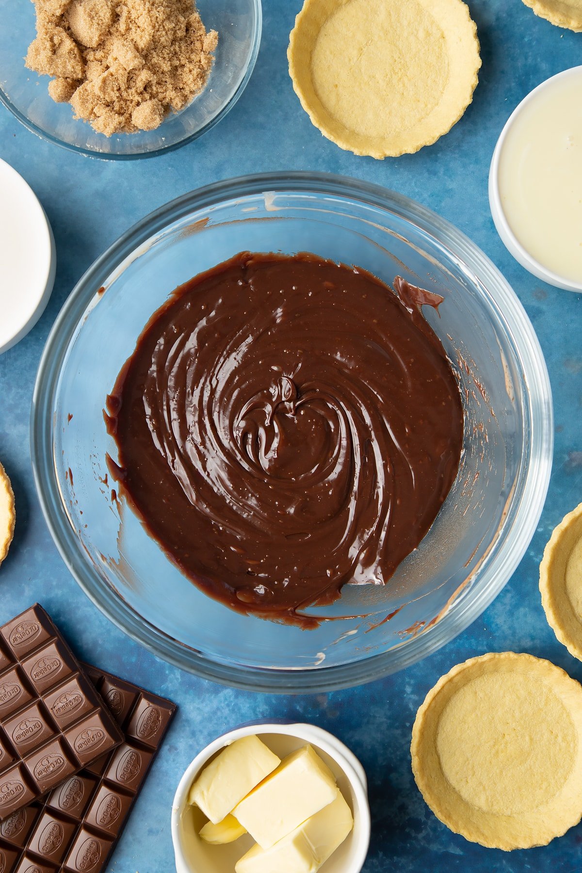 Freshly made milk chocolate ganache in a large mixing bowl. Ingredients to make mini chocolate tarts surround the bowl. 